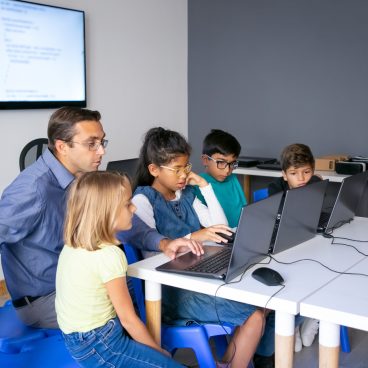 Multiethnic pupils doing task with help of teacher. Concentrated man in glasses sitting with children at table and looking at laptop screen. Childhood, communication and digital education concept